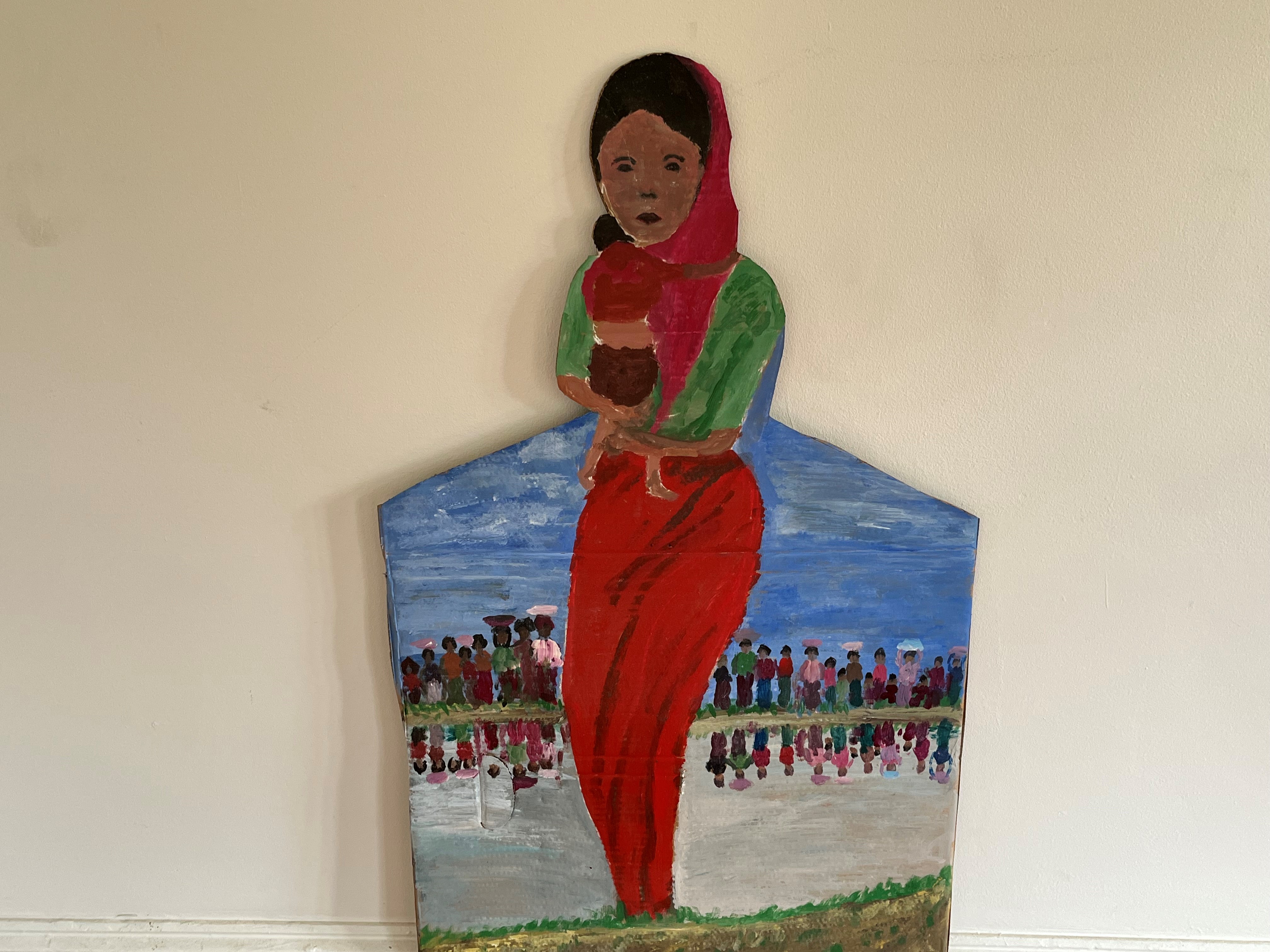 Cropped cardboard artwork, a mother wearing a loose scarf on her head hold a baby in her arms, its arm is cradled around its mothers neck and its head is pointing over her shoulder, in the background is a blue sky and many people lined up with buckets resting on their heads